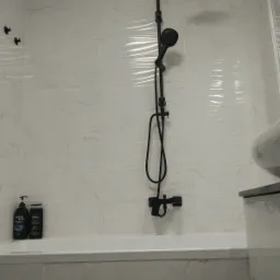 Nowoczesna łazienka z czarną armaturą: deszczownica i słuchawka prysznicowa na tle białych, marmurkowych płytek. Na wannie stoją dwa czarne pojemniki z kosmetykami.