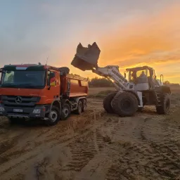 Pomarańczowa ciężarówka wywrotka i biała ładowarka Liebherr na placu budowy o zmierzchu, z łyżką ładowarki uniesioną nad skrzynią ładunkową ciężarówki.