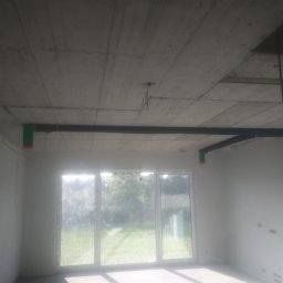 WENT4INSTAL - Surowy strop z widocznymi elementami instalacji rekuperacji, czarne rury z zielonymi zakończeniami, duże okno z widokiem na zieleń.