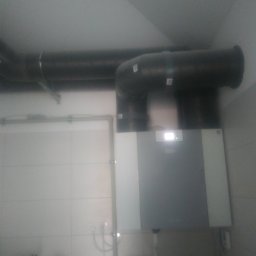 WENT4INSTAL - Szara jednostka centralna wentylacyjna z widocznym ekranem i podłączonymi czarnymi rurami wentylacyjnymi w pomieszczeniu wyłożonym białymi płytkami.