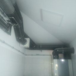 WENT4INSTAL - Szare, izolowane rury wentylacyjne pod sufitem na poddaszu, połączone z jednostką centralną na ścianie z ceramicznych płytek, widoczna instalacja elektryczna i hydrauliczna.