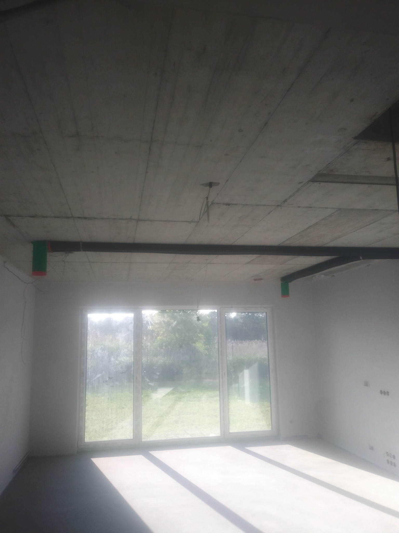 Surowy strop z widocznymi elementami instalacji rekuperacji, czarne rury z zielonymi zakończeniami, duże okno z widokiem na zieleń.