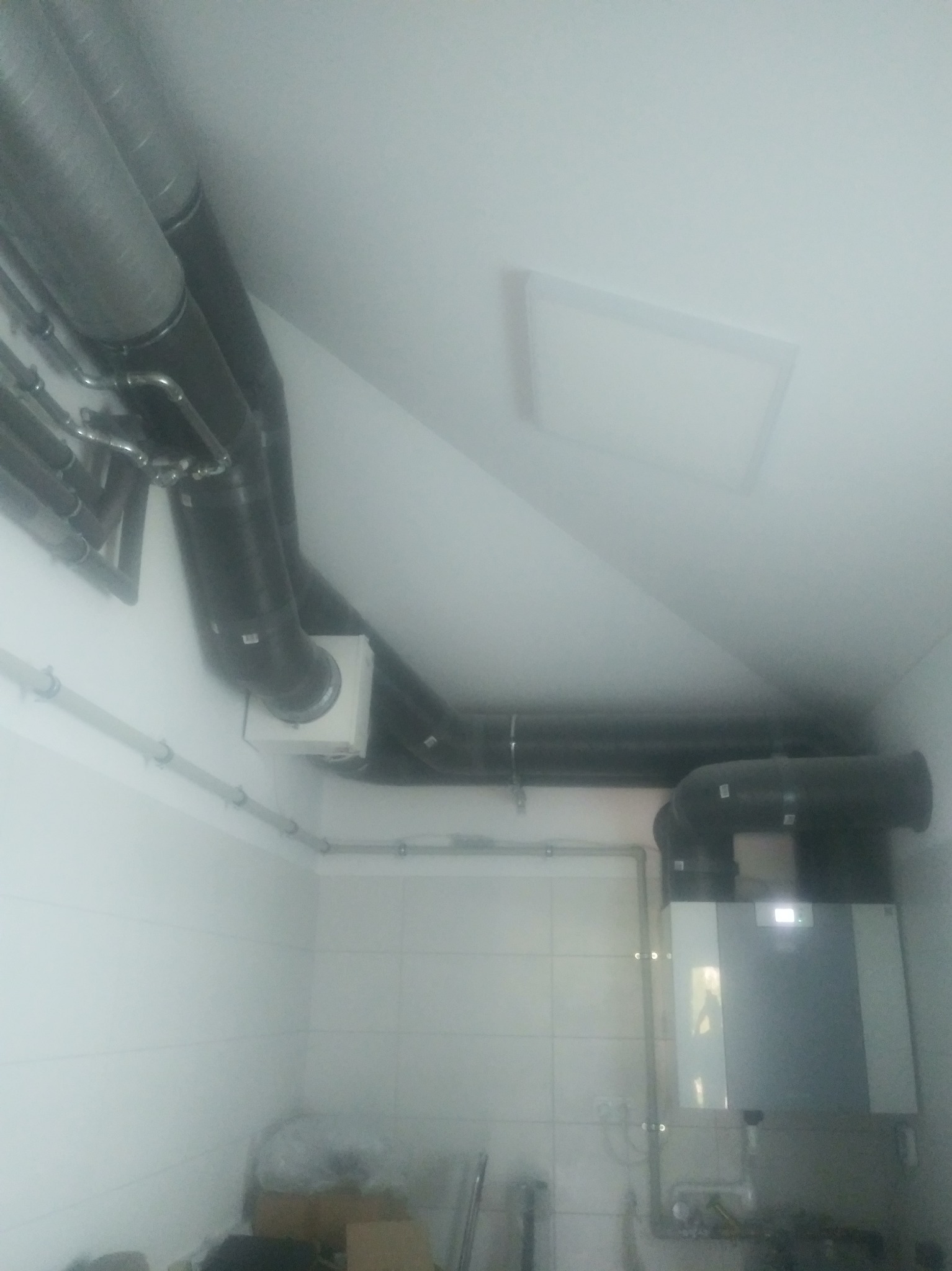 Szare, izolowane rury wentylacyjne pod sufitem na poddaszu, połączone z jednostką centralną na ścianie z ceramicznych płytek, widoczna instalacja elektryczna i hydrauliczna.