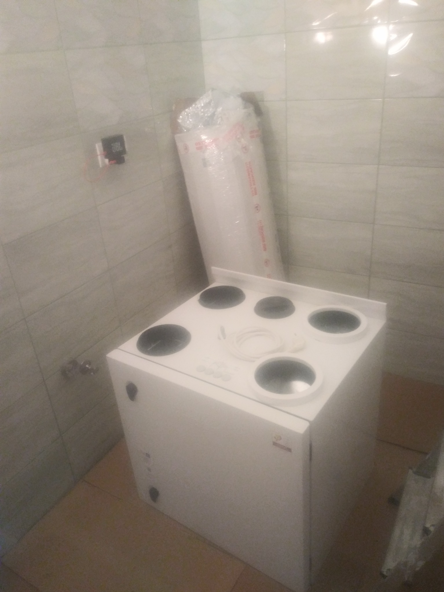 Kompaktowa centrala wentylacyjna z okrągłymi króćcami w białej obudowie, zapakowana w folię ochronną, umieszczona w rogu pomieszczenia z wykończeniem z płytek ceramicznych.