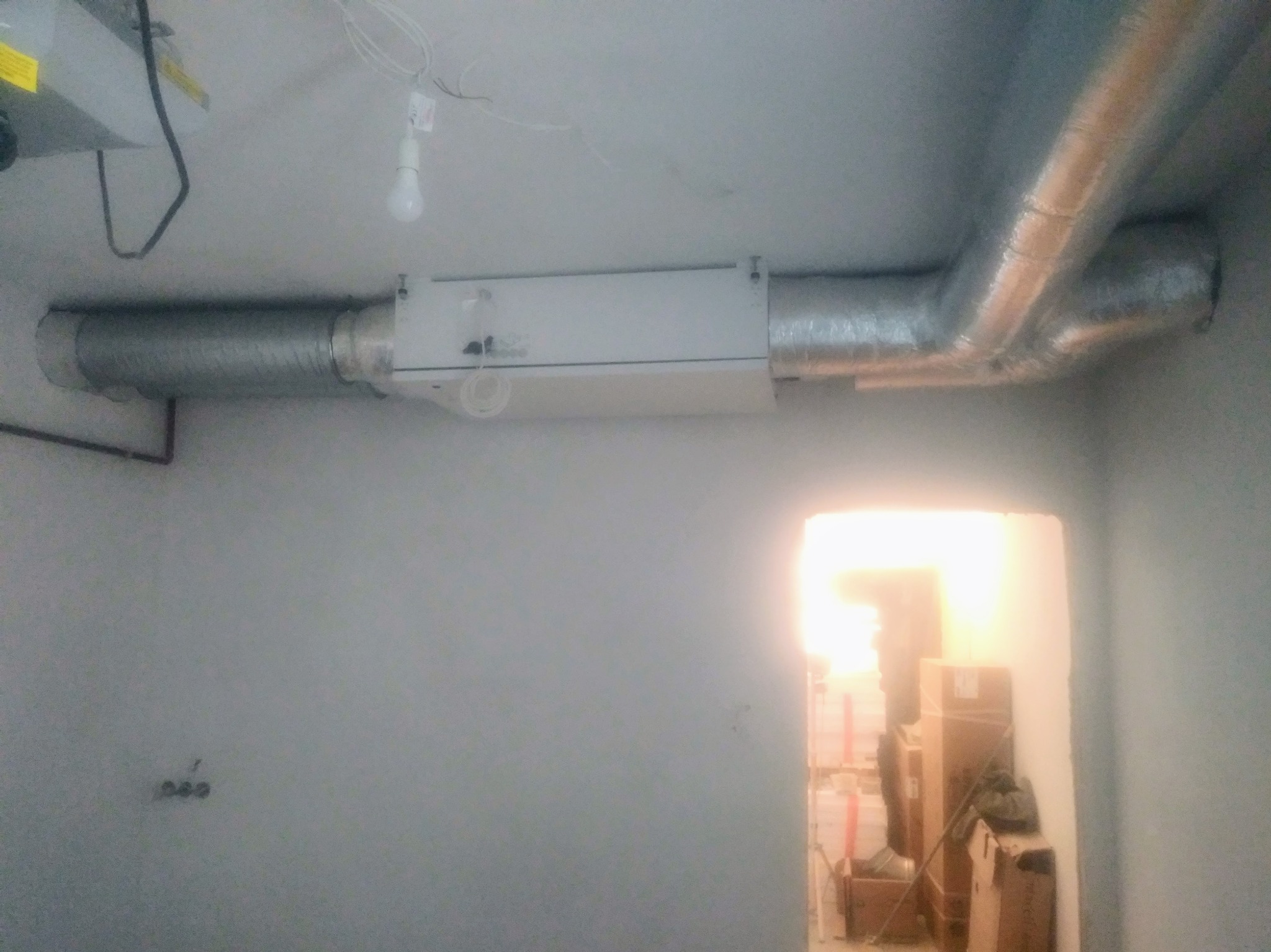 Instalacja centrali wentylacyjnej z rurami spiro na suficie w pomieszczeniu w trakcie wykańczania, widoczne kable i żarówka.