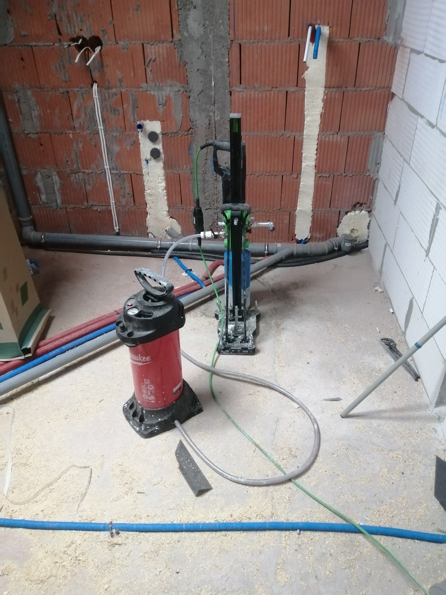 Montaż instalacji w surowym wnętrzu: czerwona pompa ciśnieniowa Milwaukee, rury instalacyjne i narzędzia na tle ceglanej ściany z wyciętymi otworami.