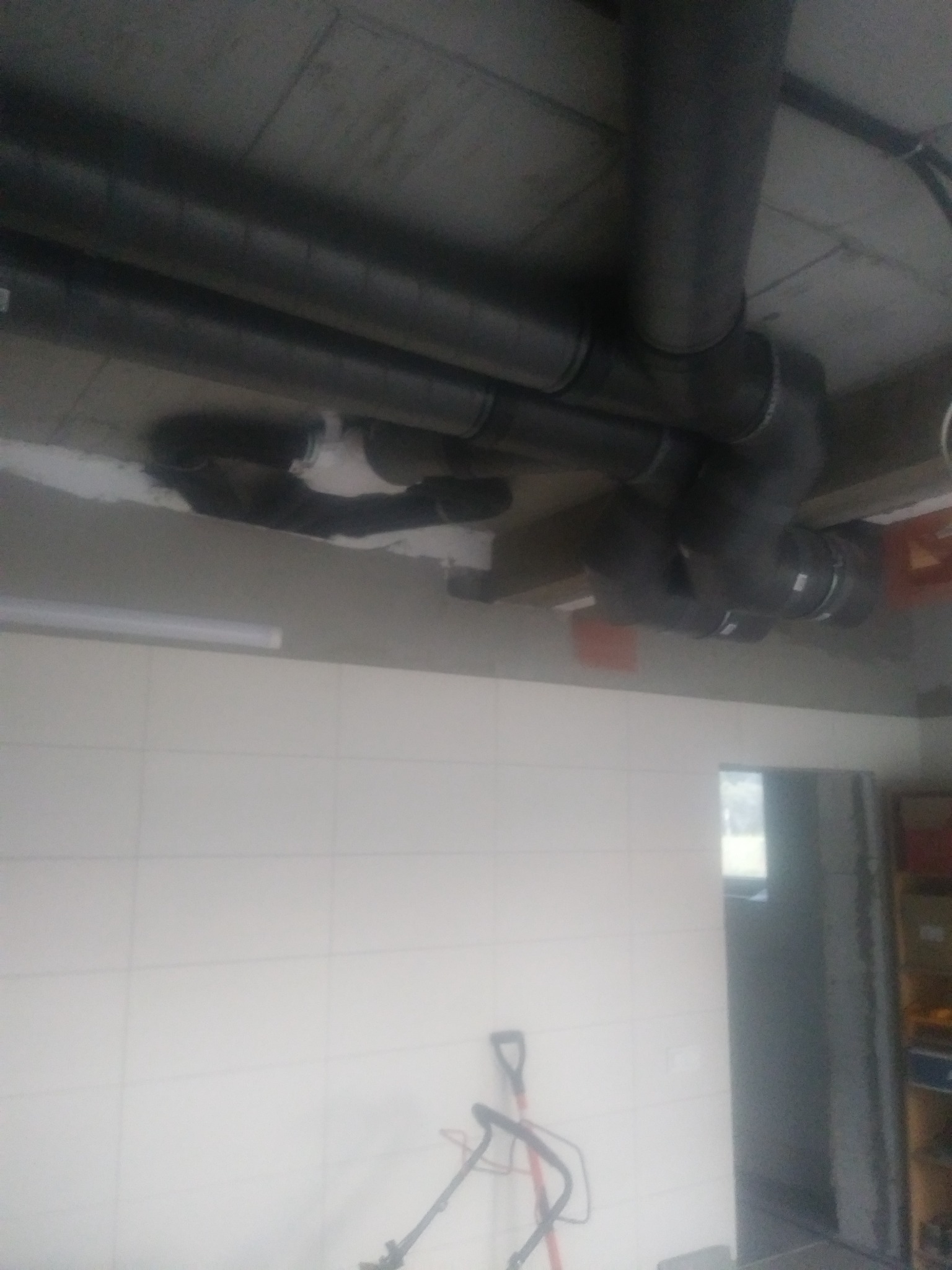 Szare rury wentylacyjne pod sufitem w pomieszczeniu z białymi płytkami. Widoczne narzędzia na podłodze. Instalacja rekuperacji w trakcie montażu.