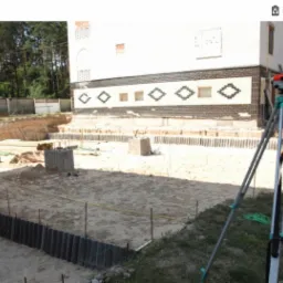 Teren budowy z wykopem, widoczne betonowe fundamenty i poziomica geodezyjna na statywie, w tle budynek z dekoracyjnym pasem z cegieł.