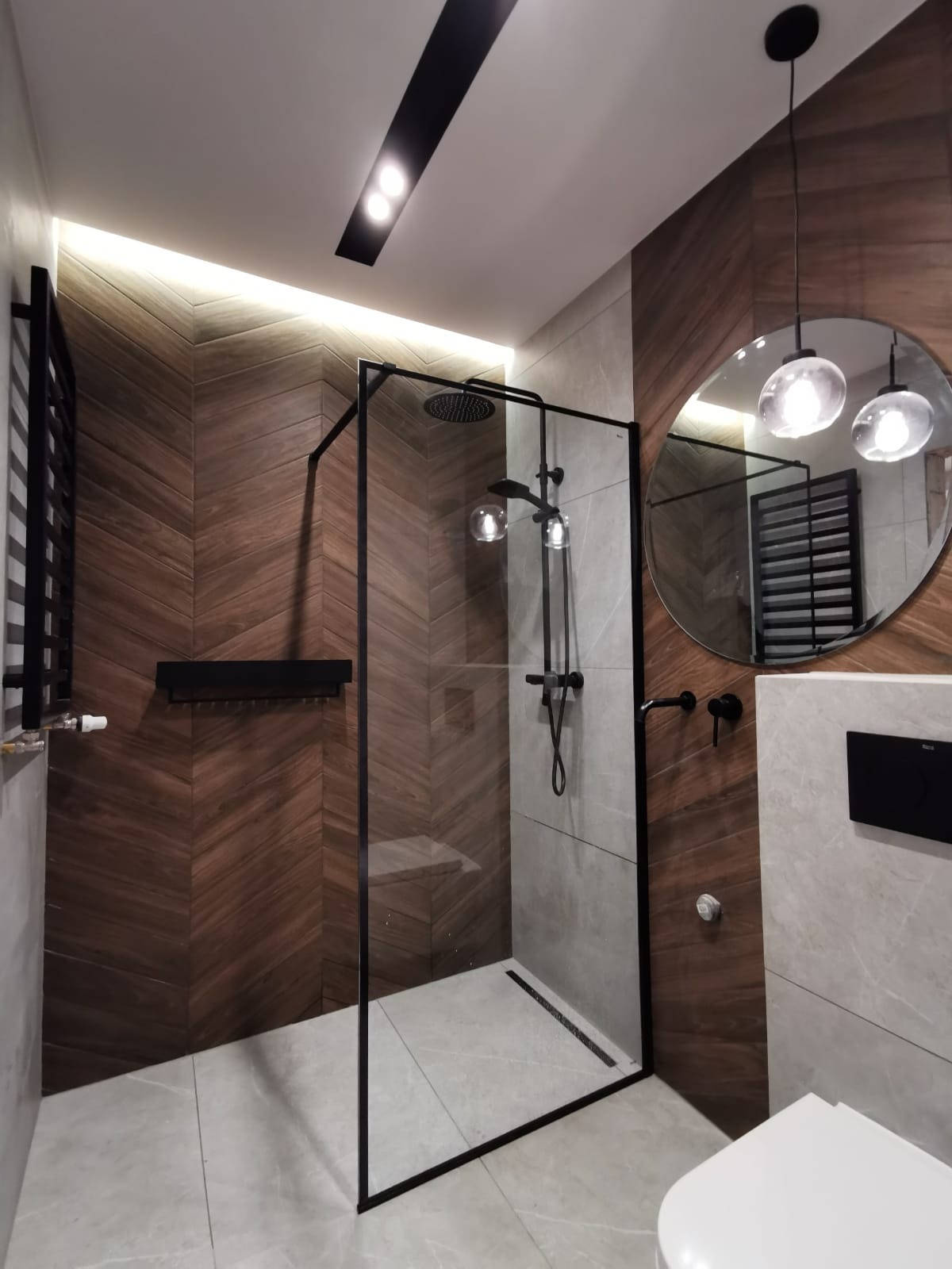 Nowoczesna łazienka z kabiną prysznicową typu walk-in z czarnymi profilami, drewnopodobne płytki ułożone w jodełkę na ścianie, okrągłe lustro z wiszącymi lampami i czarny grzejnik drabinkowy