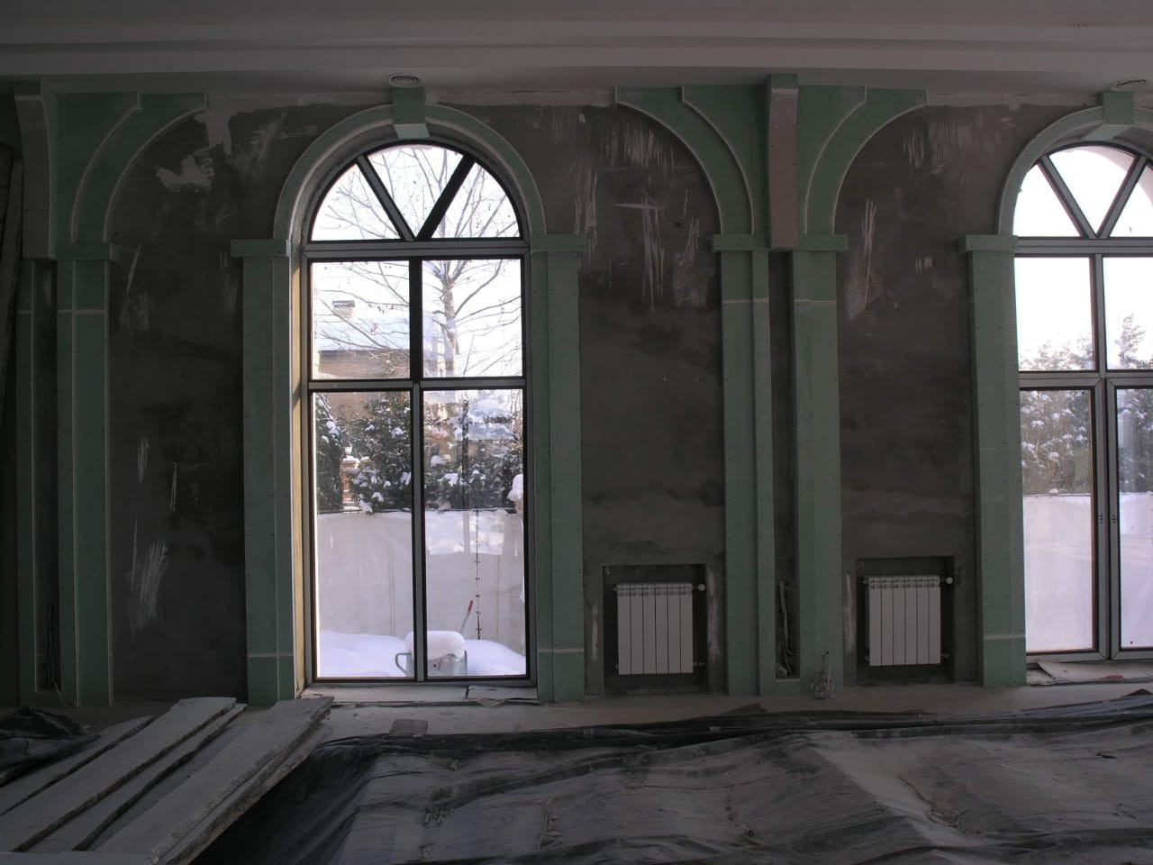 Wnętrze w trakcie prac wykończeniowych, surowa ściana z widocznymi śladami szpachlowania, zamontowane grzejniki, okna łukowe z widokiem na zaśnieżony ogród.