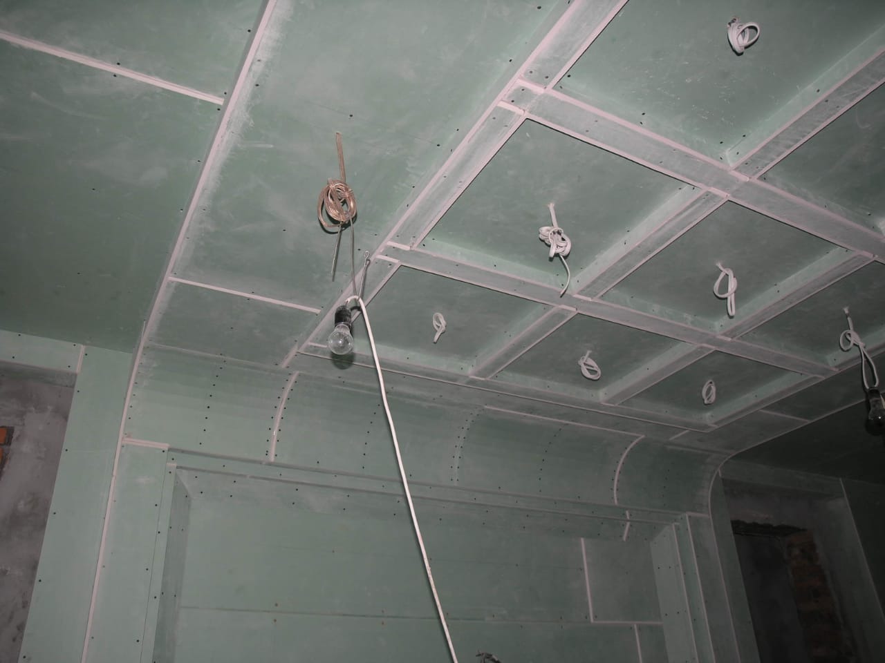 Montaż płyt gipsowo-kartonowych na suficie i ścianach, widoczne kable oświetleniowe, surowy stan wykończenia wnętrza.