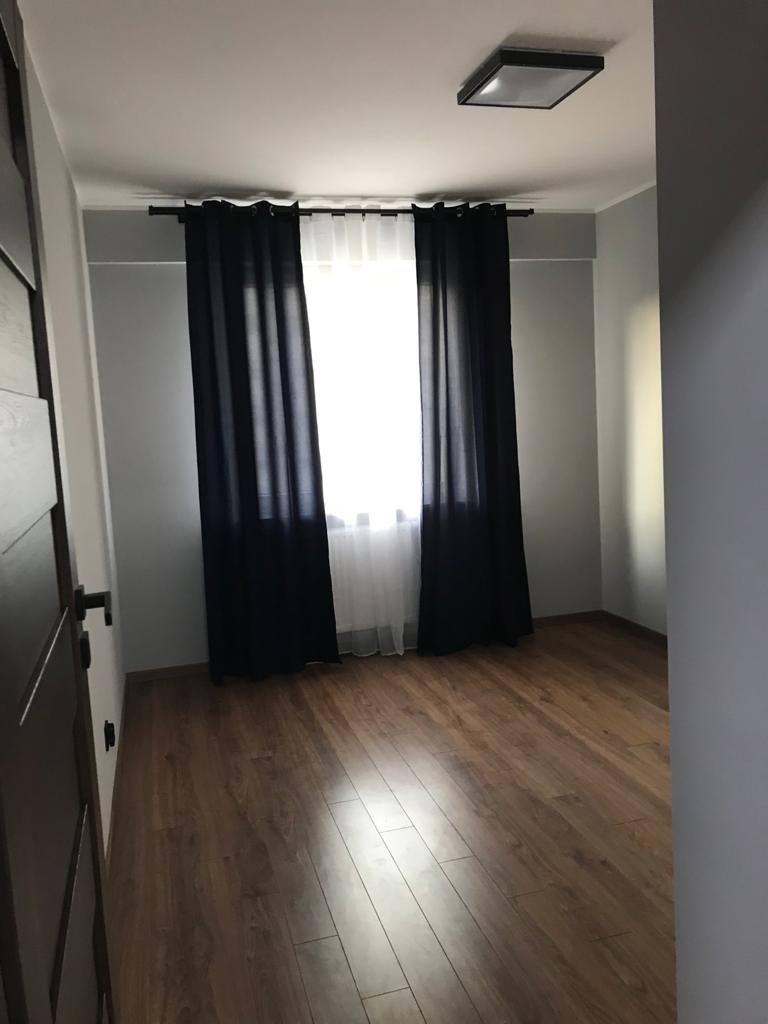 Pusty pokój z drewnianą podłogą, czarnymi zasłonami i białymi firankami, po remoncie, z widocznym oświetleniem sufitowym.