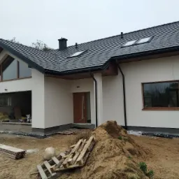 Nowoczesny dom jednorodzinny z ocieploną, białą elewacją, ciemną dachówką i drewnianymi oknami, widok z zewnątrz na plac budowy z stertą piasku i paletami.