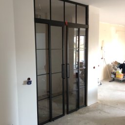 Firma SOW-STAL - Czarno-ramowe, przeszklone drzwi przesuwne w stylu loftowym, zamontowane w surowym wnętrzu w trakcie prac wykończeniowych, widoczne ślady farby na podłodze i niezabezpieczone gniazdka elektryczne.