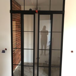 Firma SOW-STAL - Dwuskrzydłowe, przeszklone drzwi metalowe w stylu loftowym, z czarnymi szprosami, widoczne na tle surowej cegły i białych ścian w trakcie wykończenia.