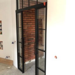 Firma SOW-STAL - Czarne, przeszklone drzwi przesuwne z metalową ramą w industrialnym stylu, z widoczną ścianą z czerwonej cegły w tle, w surowym wnętrzu podczas remontu.