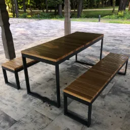 Zestaw mebli ogrodowych: stół z drewnianym blatem i metalową ramą oraz dwie ławki, ustawiony na kamiennej posadzce pod drewnianą altaną, w tle widoczny park.