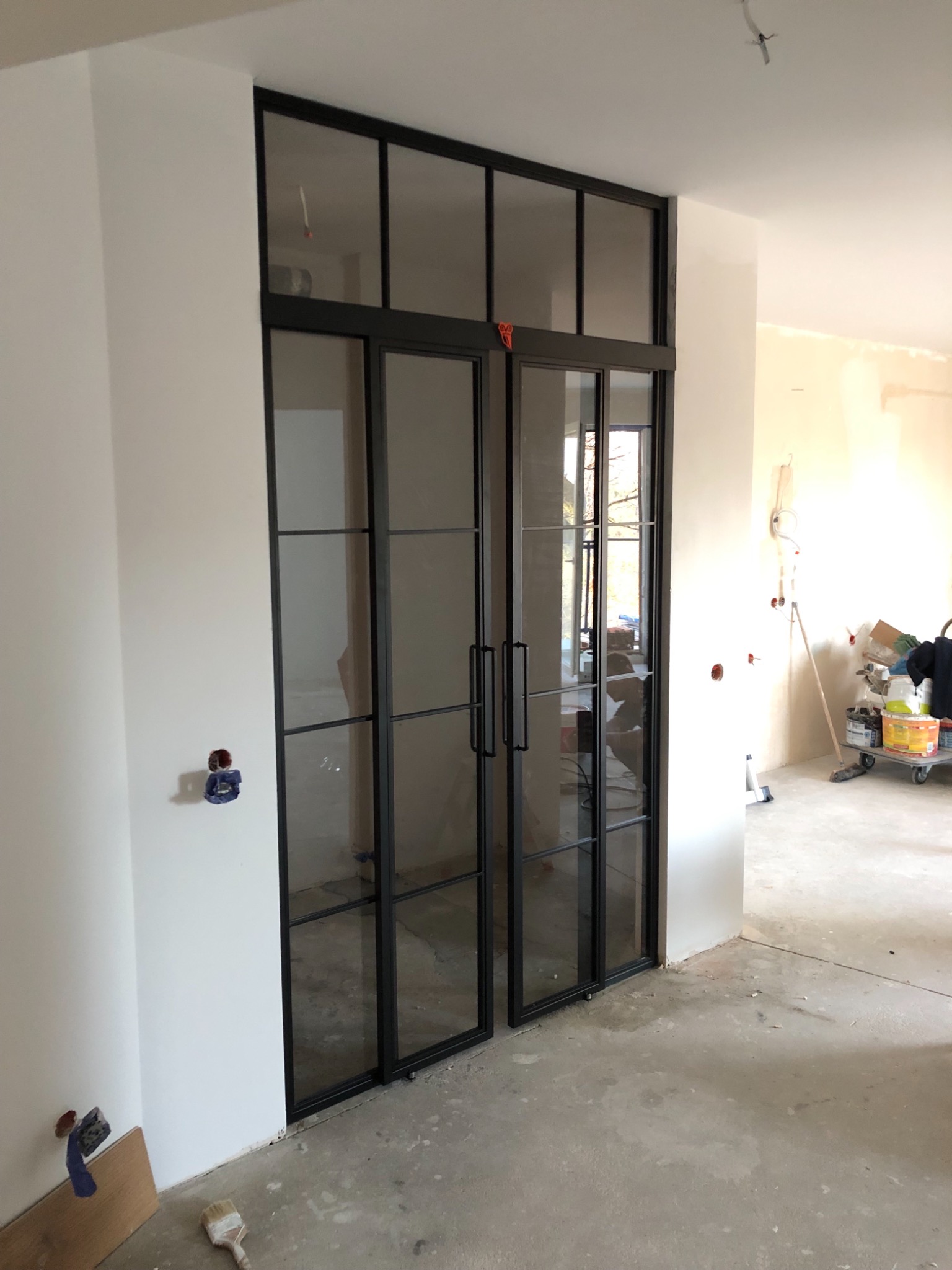 Czarno-ramowe, przeszklone drzwi przesuwne w stylu loftowym, zamontowane w surowym wnętrzu w trakcie prac wykończeniowych, widoczne ślady farby na podłodze i niezabezpieczone gniazdka elektryczne.