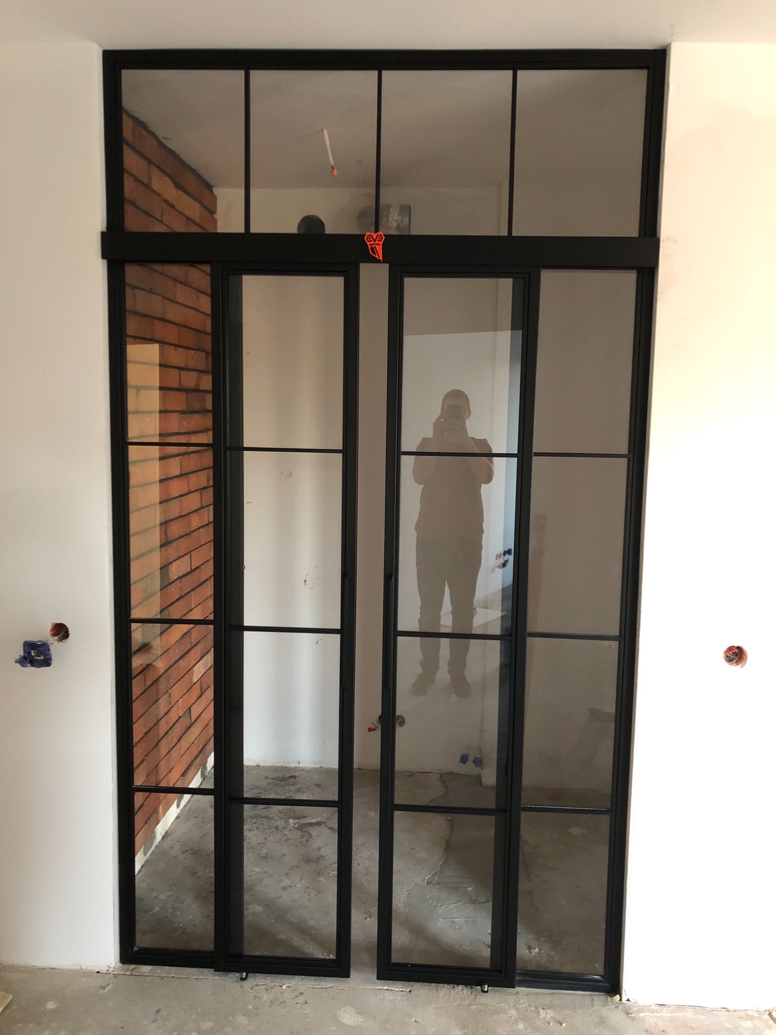 Dwuskrzydłowe, przeszklone drzwi metalowe w stylu loftowym, z czarnymi szprosami, widoczne na tle surowej cegły i białych ścian w trakcie wykończenia.
