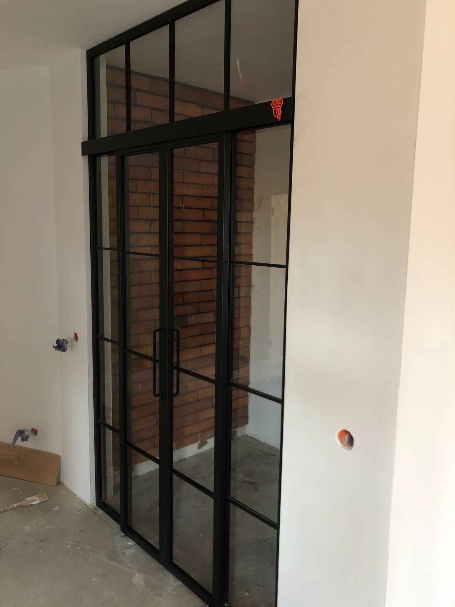 Loftowe drzwi przesuwne z czarnymi szprosami i szybami, widok na ceglaną ścianę, surowy beton na podłodze, trwają prace wykończeniowe.