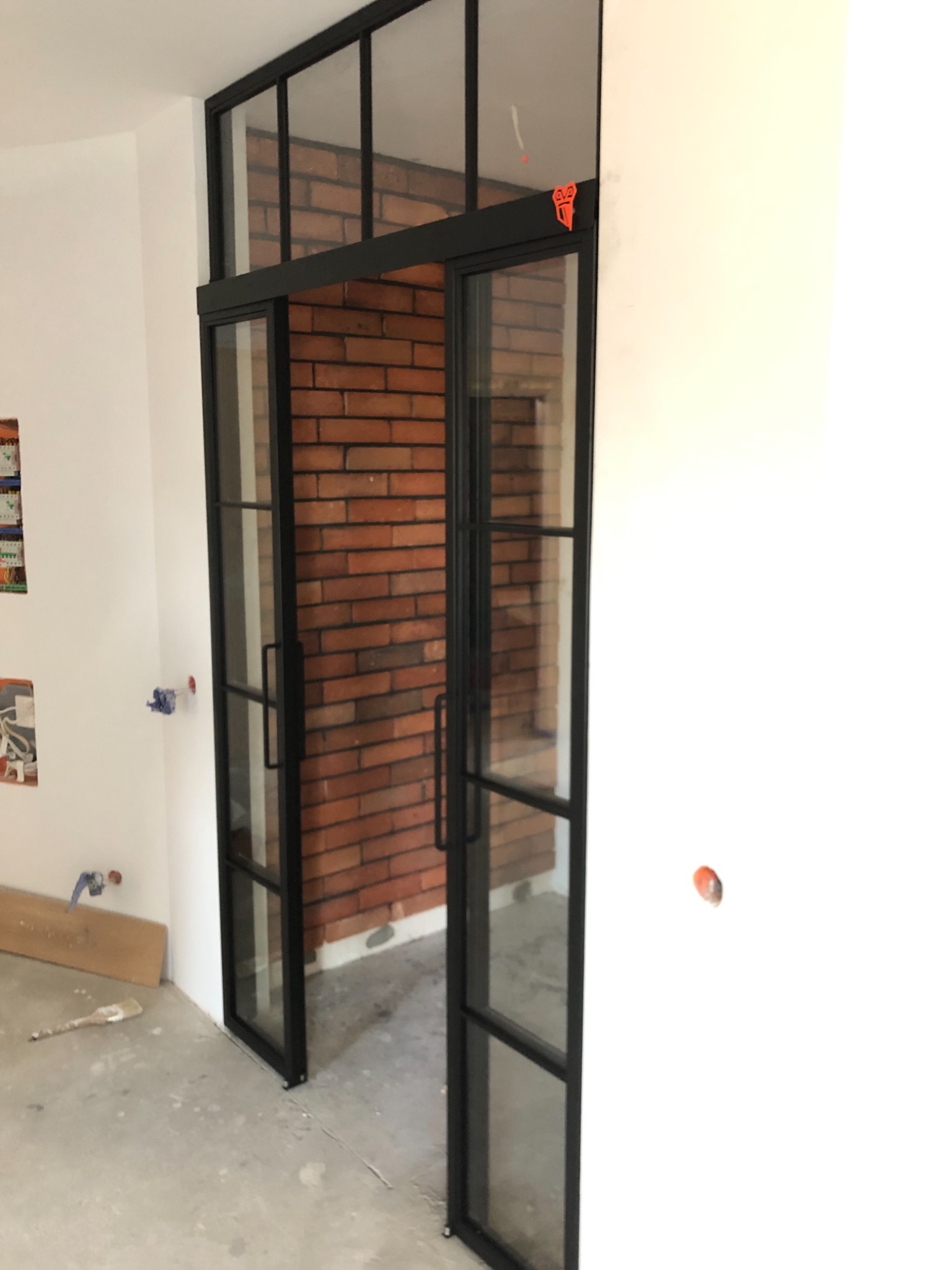 Czarne, przeszklone drzwi przesuwne z metalową ramą w industrialnym stylu, z widoczną ścianą z czerwonej cegły w tle, w surowym wnętrzu podczas remontu.