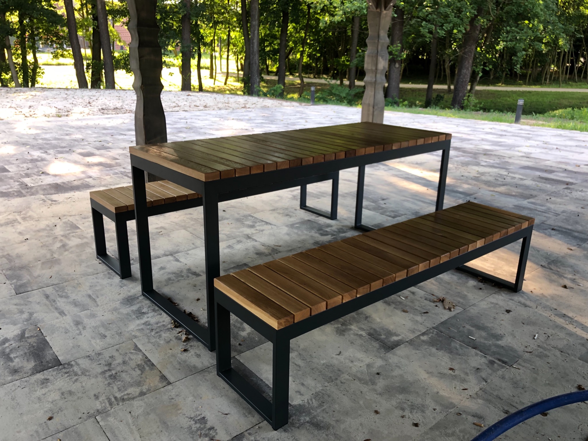 Zestaw mebli ogrodowych: stół z drewnianym blatem i metalową ramą oraz dwie ławki, ustawione na kamiennej posadzce w zacienionym ogrodzie.