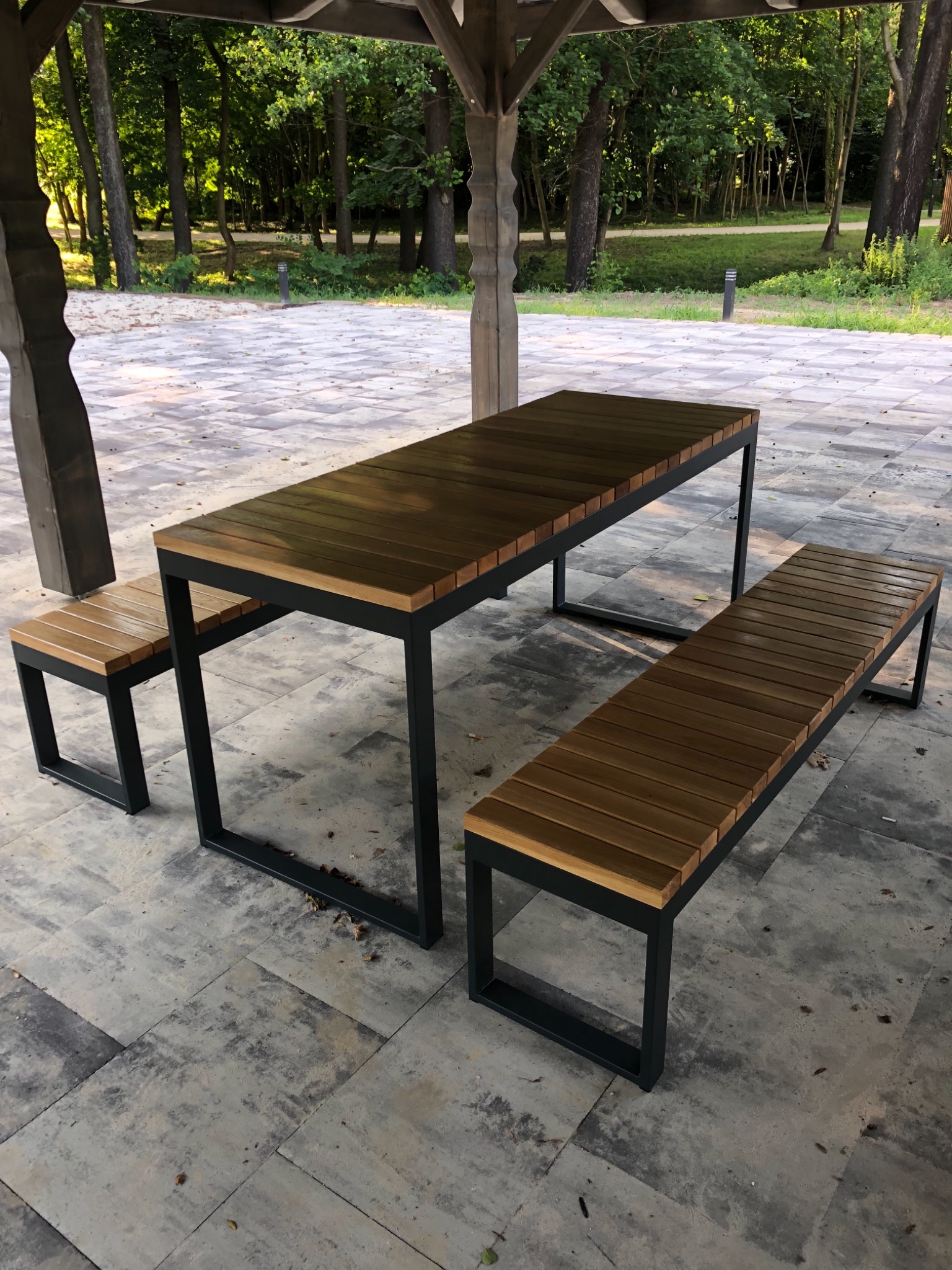 Zestaw mebli ogrodowych: stół z drewnianym blatem i metalową ramą oraz dwie ławki, ustawiony na kamiennej posadzce pod drewnianą altaną, w tle widoczny park.
