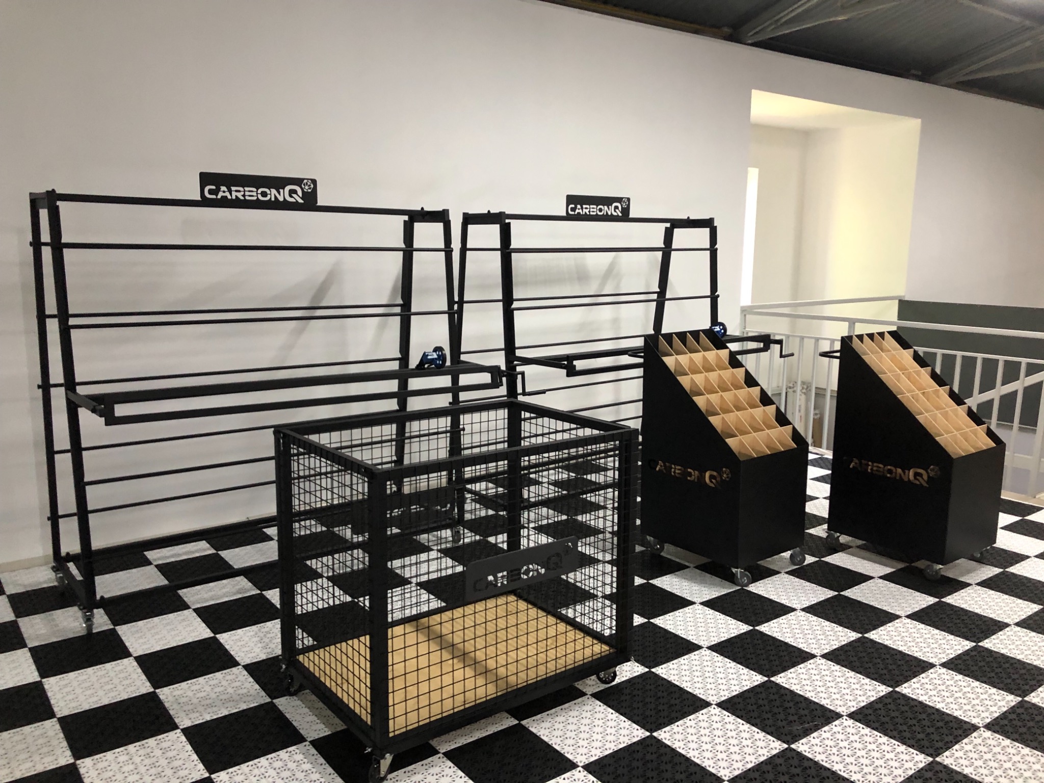 Metalowe konstrukcje na kółkach z logo CarbonQ: regał z poziomymi prętami, klatka z drewnianym dnem oraz dwa pojemniki z przegródkami, ustawione na czarno-białej podłodze w szachownicę.