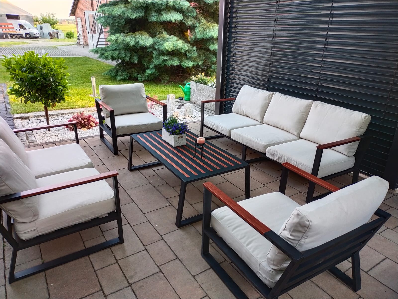 Zestaw mebli ogrodowych z metalową ramą i jasnymi poduchami na brukowanym tarasie, z widokiem na zadbany ogród i rolety okienne.
