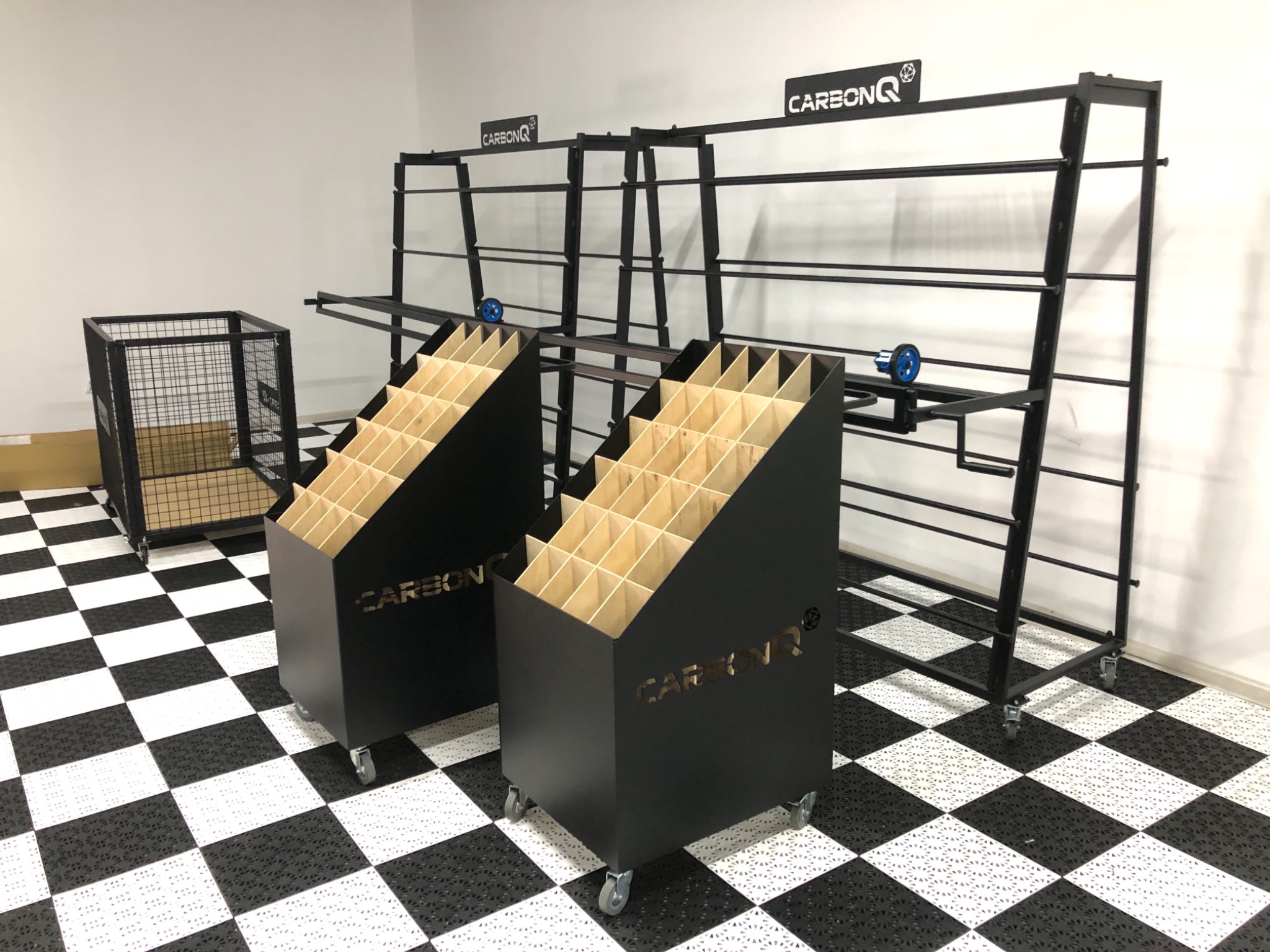 Metalowe regały warsztatowe z półkami i kosz na kółkach w pomieszczeniu z podłogą w szachownicę.