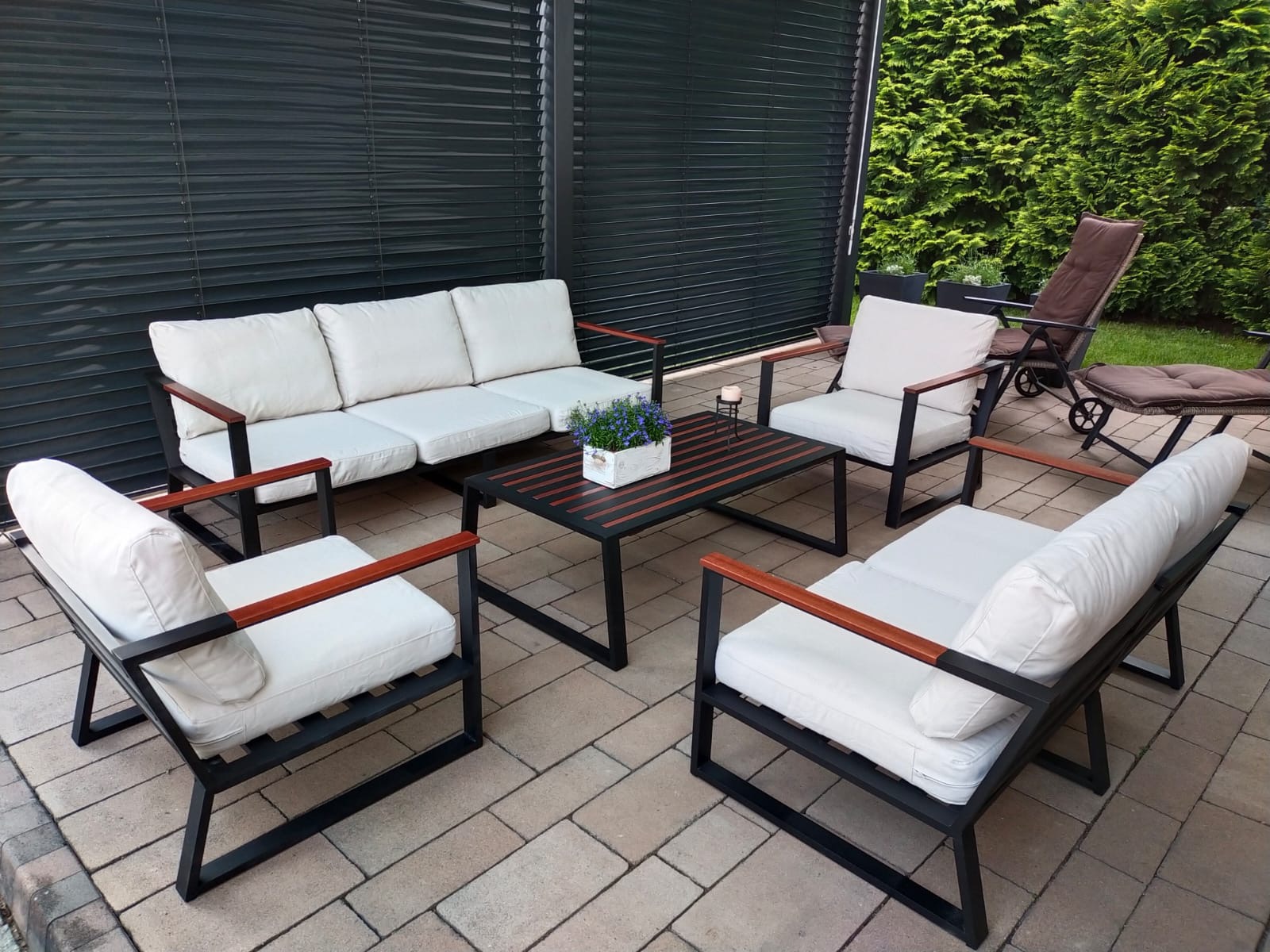 Zestaw mebli ogrodowych z czarną metalową ramą, drewnianymi podłokietnikami i białymi poduszkami, ustawiony na tarasie z kostki brukowej, z widokiem na ciemne rolety i zielone tuje.