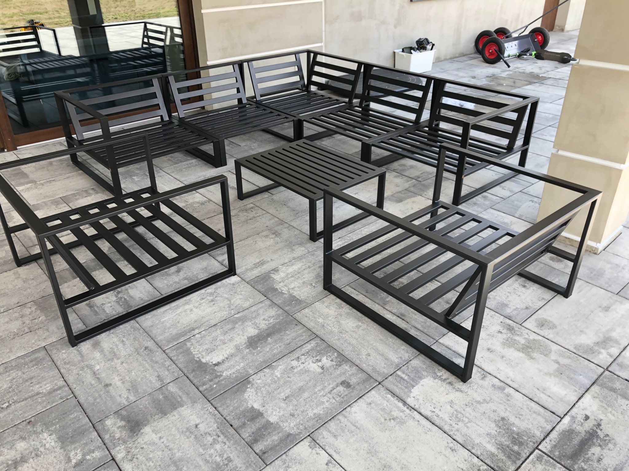 Komplet metalowych mebli ogrodowych w kolorze czarnym, składający się z sofy narożnej, dwóch foteli i stolika kawowego, ustawiony na szarym brukowanym patio przy domu.