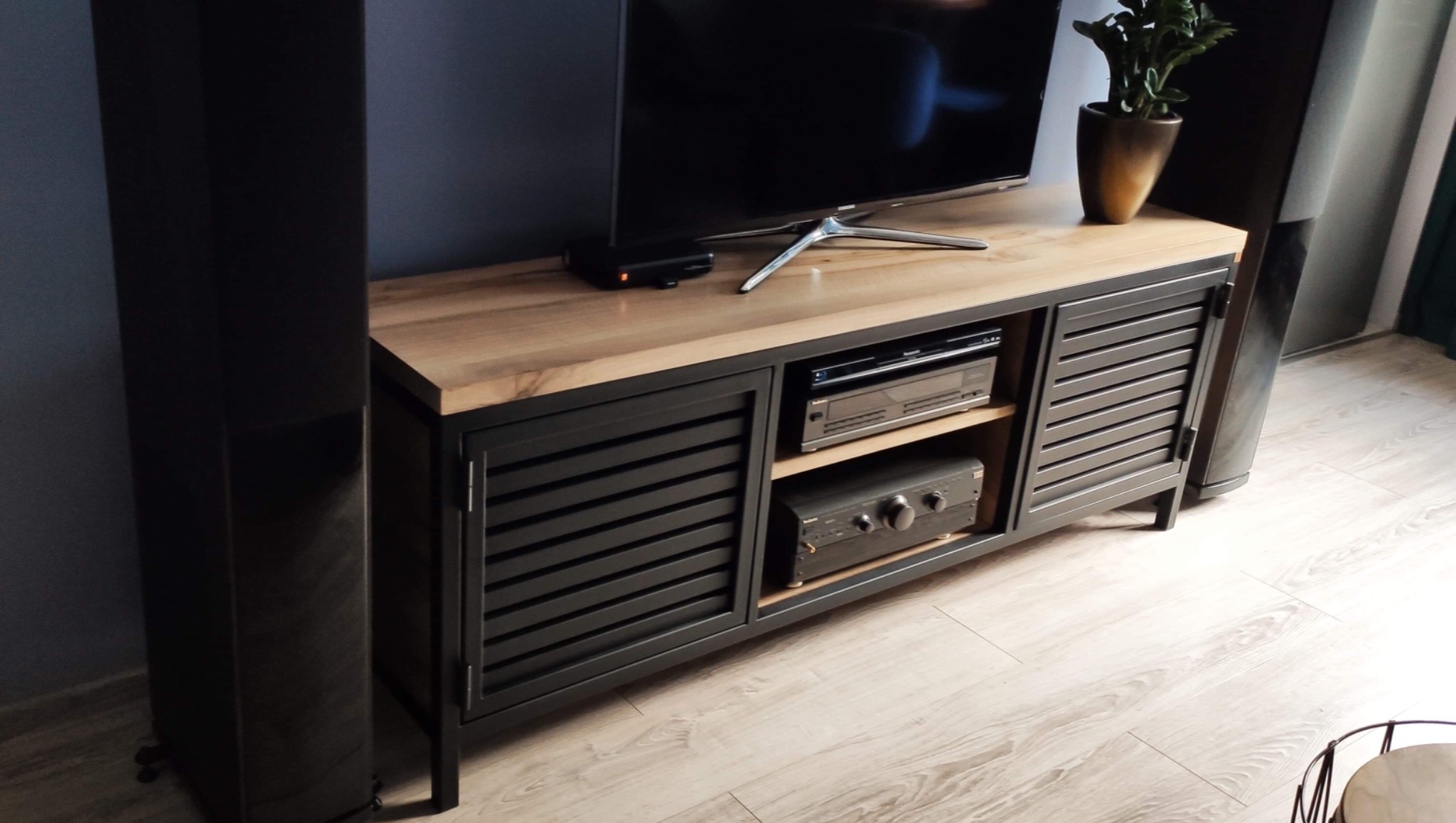 Szafka RTV w stylu industrialnym z drewnianym blatem i metalowymi, żaluzjowymi drzwiczkami, na której stoi telewizor, dekoder i roślina doniczkowa.
