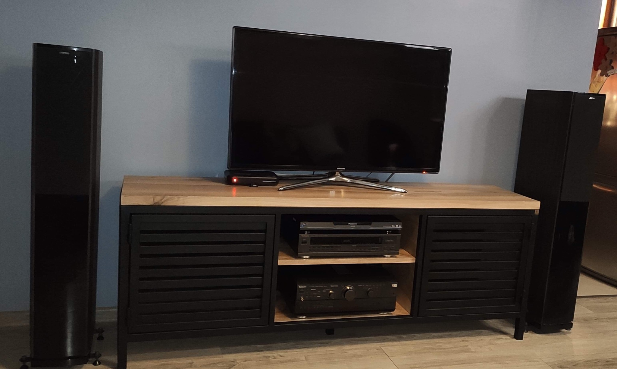 Czarna szafka RTV z drewnianym blatem, zamykana żaluzjowymi drzwiczkami, na której stoi telewizor, a w środku sprzęt audio; obok stoją czarne kolumny głośnikowe.