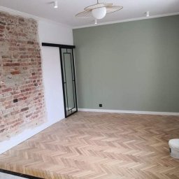 Wykończenia A-Z - Pokój po renowacji z odsłoniętym fragmentem ceglanej ściany, pomalowaną na oliwkowo ścianą, parkietem ułożonym w jodełkę i przesuwnymi drzwiami z czarną ramą. Widoczny również biały pojemnik na farbę.