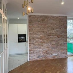 Wykończenia A-Z - Salon z parkietem w jodełkę, ścianą z cegły dekoracyjnej i widokiem na kuchnię z białymi meblami oraz przeszkloną szafą.