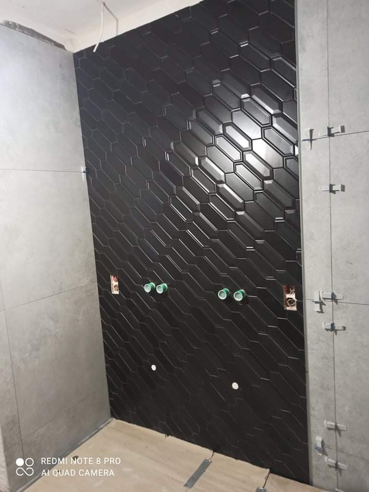 Ściana w łazience wyłożona czarnymi, błyszczącymi płytkami w kształcie wydłużonych sześciokątów. Widoczne białe punkty fugowania i wystające rury instalacyjne. Po prawej stronie ściana wyłożona...
