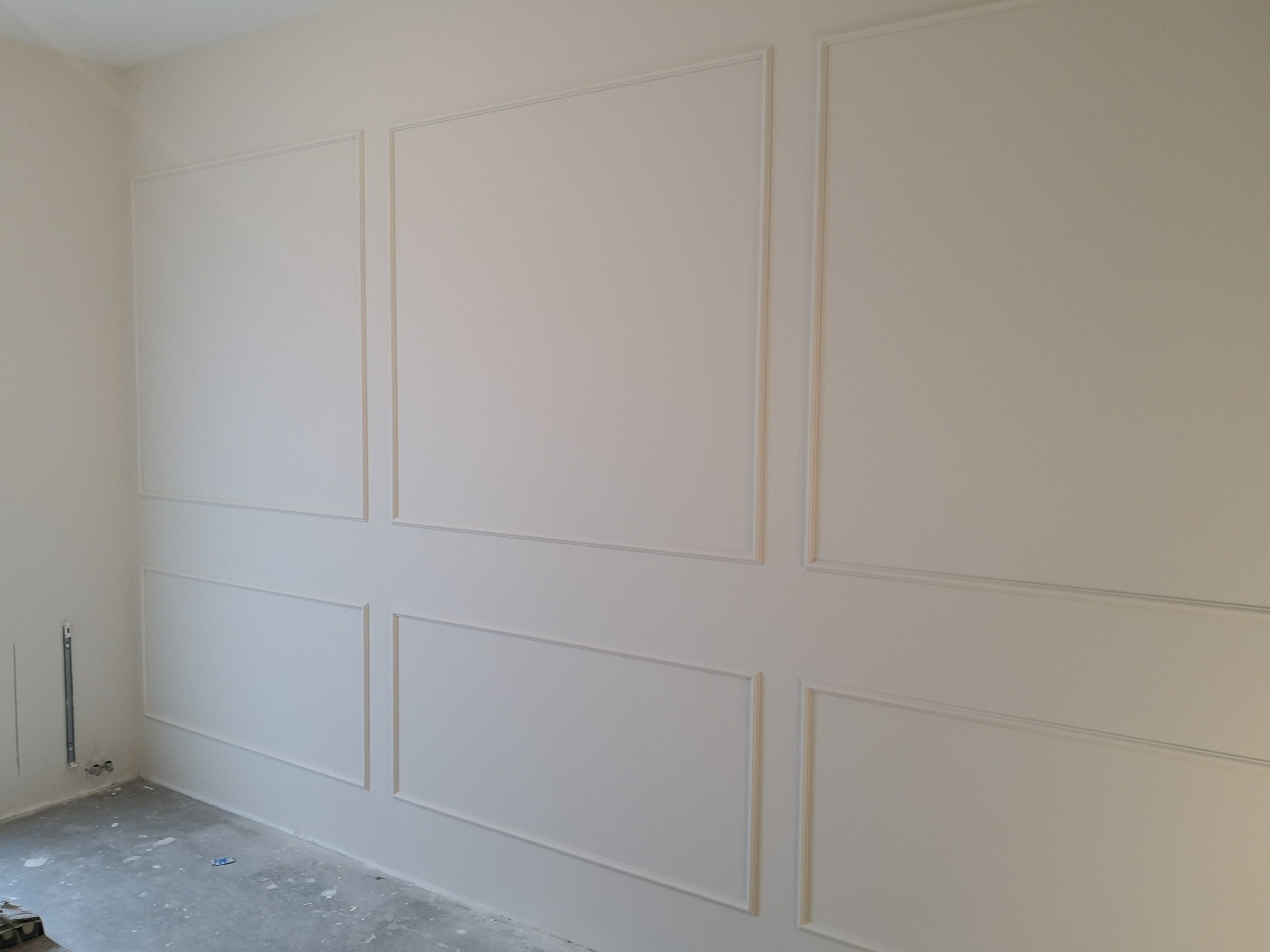 Ściana z dekoracyjnymi listwami w kształcie prostokątów, pomalowana na jasny, kremowy kolor; widoczny fragment surowej podłogi.