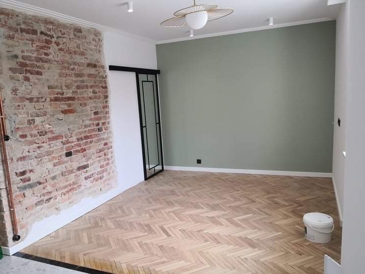 Pokój po renowacji z odsłoniętym fragmentem ceglanej ściany, pomalowaną na oliwkowo ścianą, parkietem ułożonym w jodełkę i przesuwnymi drzwiami z czarną ramą. Widoczny również biały pojemnik na farbę.