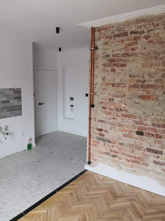 Wnętrze w trakcie prac wykończeniowych: połączenie drewnianego parkietu w jodełkę z szarą posadzką, fragment ściany z surowej czerwonej cegły z miedzianymi rurami, białe ściany i drzwi w tle...