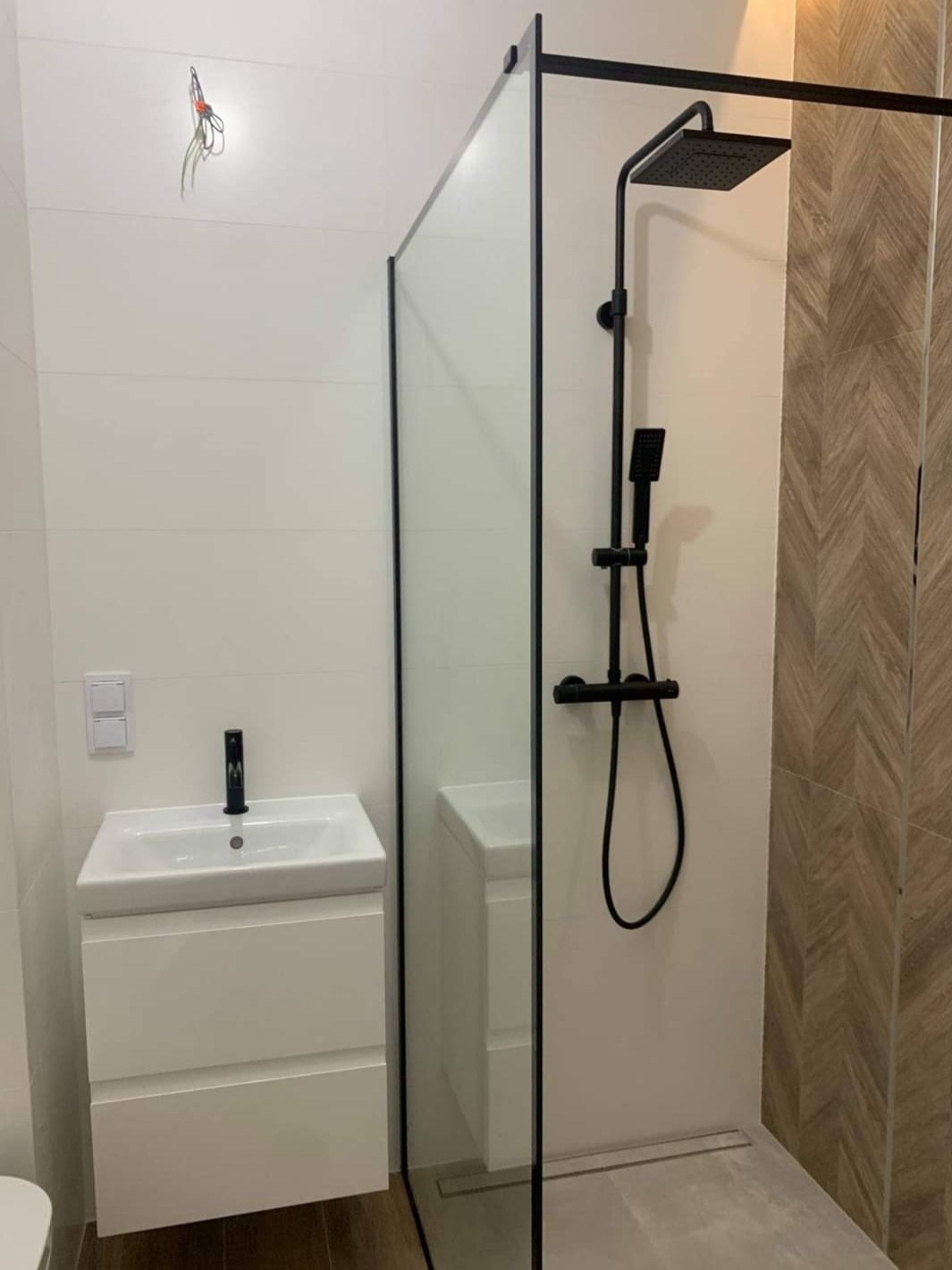 Nowoczesna łazienka z kabiną prysznicową typu walk-in z czarnymi profilami, białą szafką z umywalką i armaturą w kolorze czarnym, płytki imitujące drewno na ścianie oraz wystające kable ze ściany.