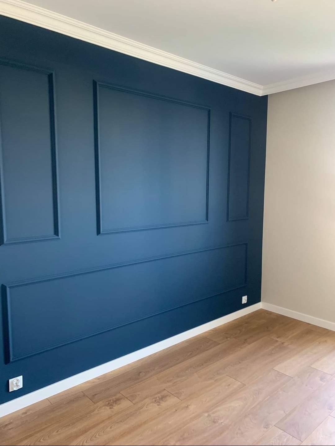 Granatowa ściana z dekoracyjnymi panelami w stylu angielskim, białe listwy przypodłogowe i drewnopodobna podłoga, widoczne gniazdka elektryczne.