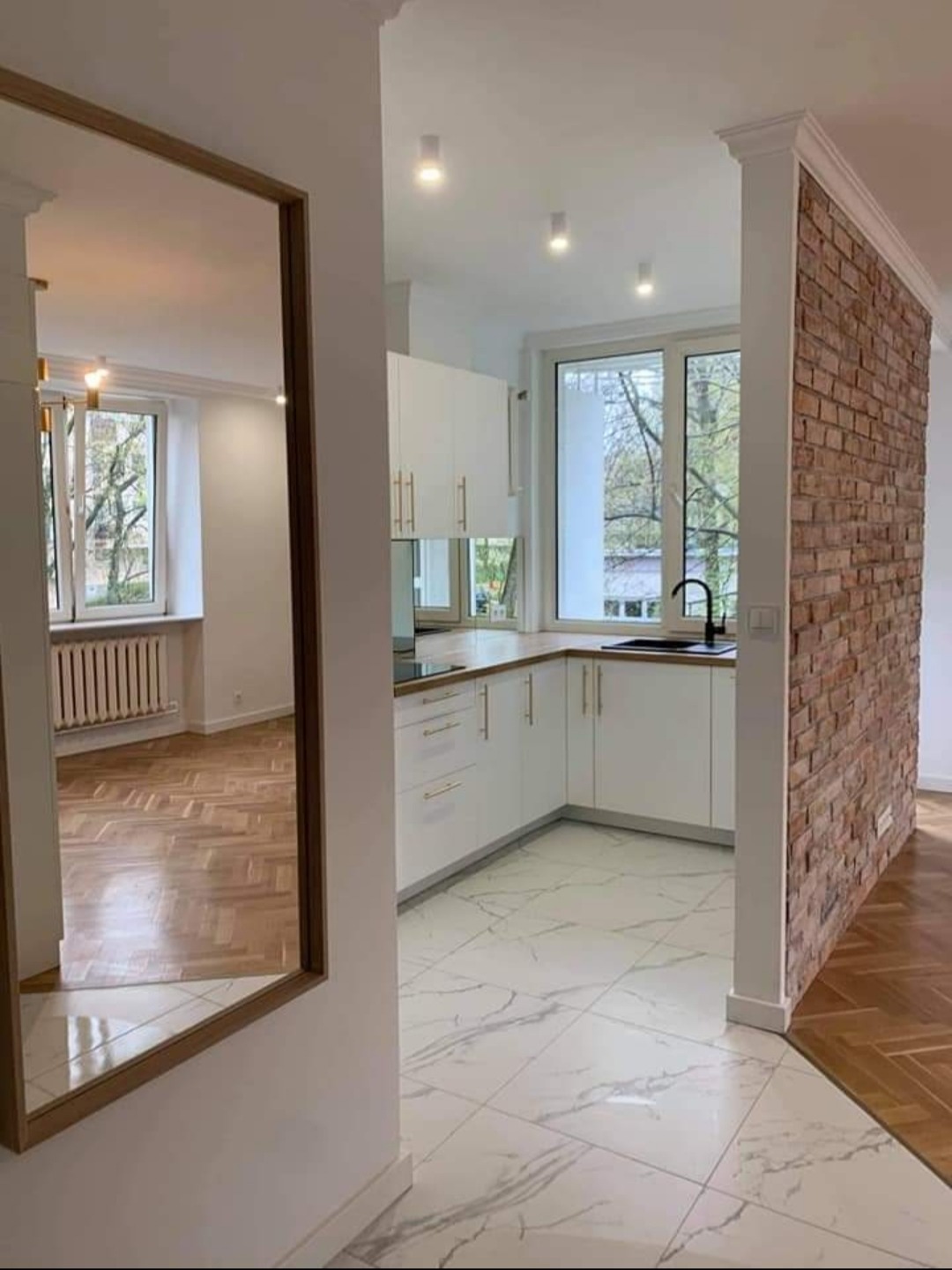 Nowoczesna kuchnia z białymi szafkami, marmurową podłogą i ścianą z cegły, widoczna przez lustro odbijające parkiet w jodełkę w sąsiednim pomieszczeniu.