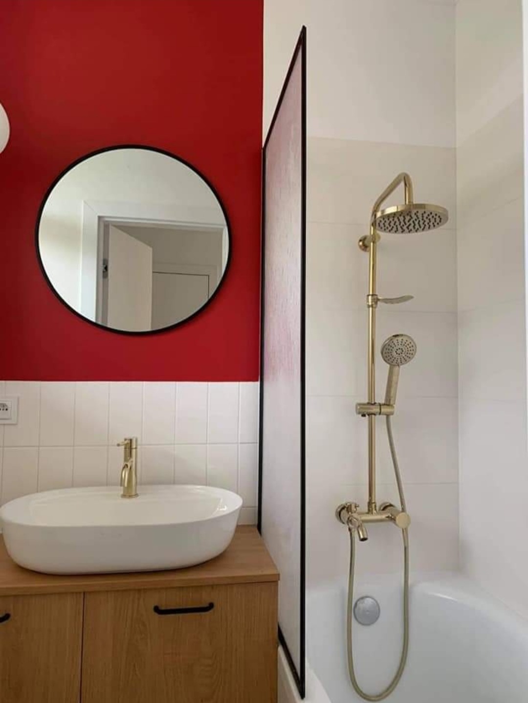 Nowoczesna łazienka z czerwonym akcentem na ścianie, okrągłe lustro z czarną ramą nad białą umywalką nablatową na drewnianej szafce, złota armatura i szklana ścianka prysznicowa z czarnym...