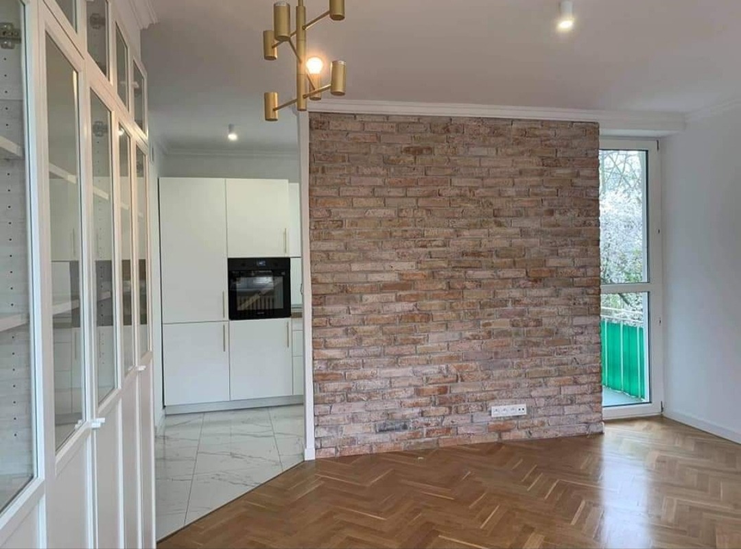 Salon z parkietem w jodełkę, ścianą z cegły dekoracyjnej i widokiem na kuchnię z białymi meblami oraz przeszkloną szafą.