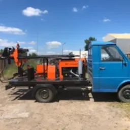 Niebieski samochód dostawczy z zamontowaną na platformie wiertnicą koloru pomarańczowego na tle błękitnego nieba i zieleni drzew.