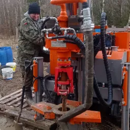 Pracownik w kamuflażu obsługuje pomarańczowo-czerwoną wiertnicę studni głębinowej, w tle las i błotniste podłoże.