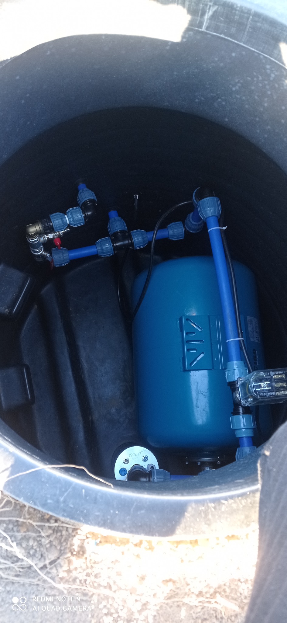 Niebieski zbiornik hydroforowy z podłączeniami w czarnej studzience, instalacja wykonana w Manowie.