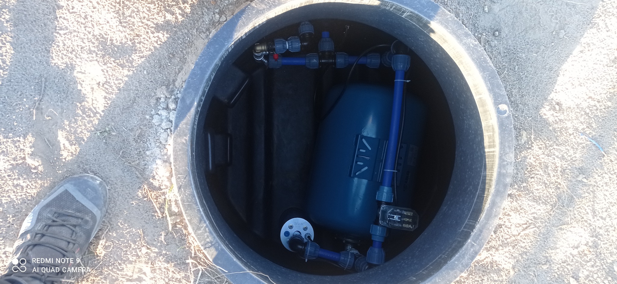 Wnętrze betonowej studni z zainstalowanym zbiornikiem hydroforowym, widoczne niebieskie rury i złączki, szara stopa buta w lewym dolnym rogu kadru.