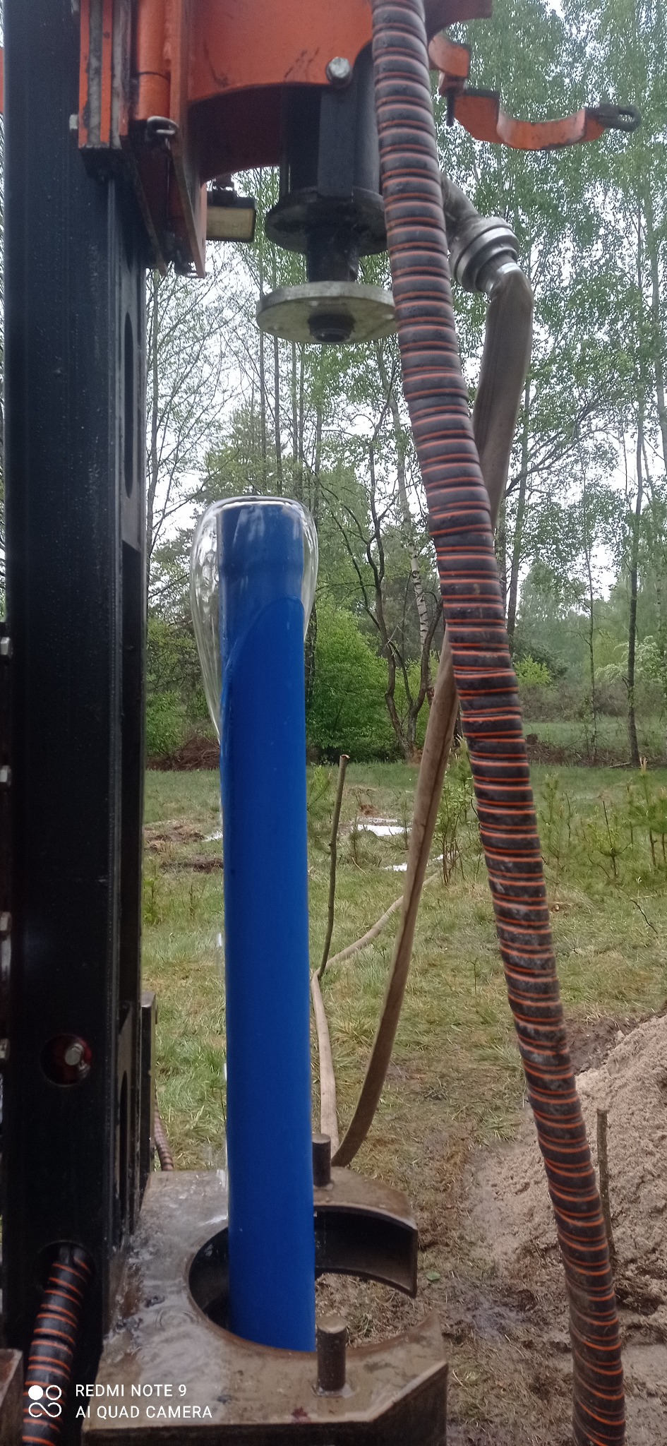Wiertnica podczas instalacji niebieskiej rury w ziemi na tle zalesionego terenu, widoczny fragment pomarańczowego węża spiralnego i czarnej konstrukcji wiertniczej.